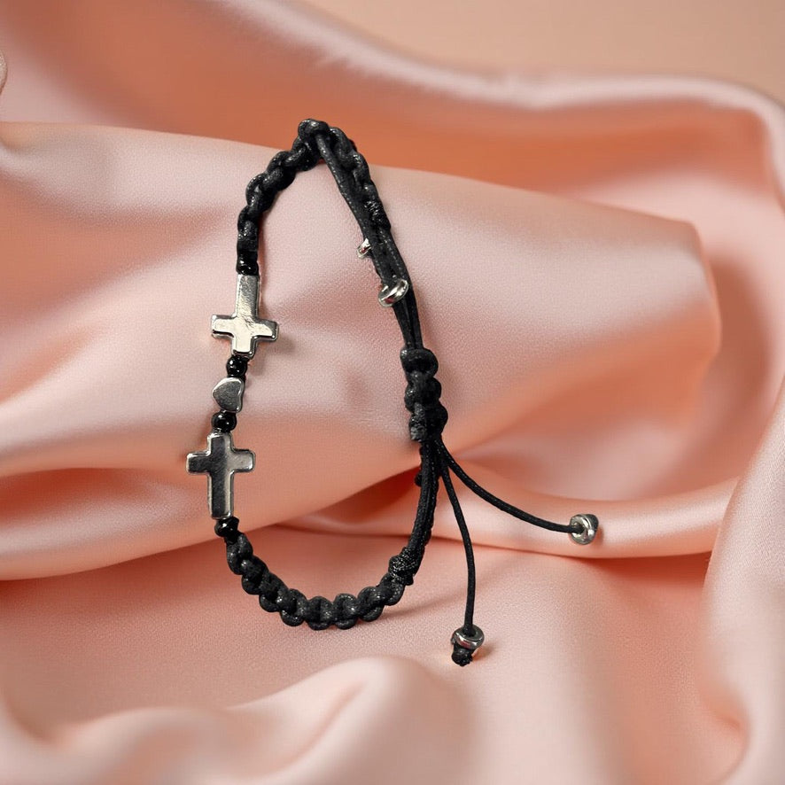 Black braided bracelet with silver cross beads on a soft pink fabric background